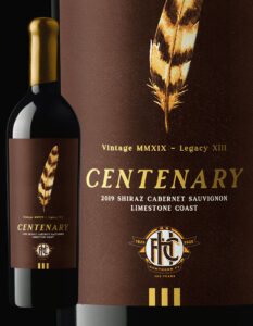 Close-up of a wine bottle labeled 'Centenary 2019 Shiraz Cabernet Sauvignon Limestone Coast.' The label is dark brown with gold text, featuring an illustrated brown-and-gold feather above the word 'Centenary' and the Hawthorn Football Club centenary logo (1925–2025) at the bottom. The bottle has a gold foil capsule and the design includes both a full bottle view and a zoomed-in section of the label.